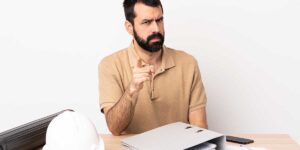 An angry builder points his finger at a desk with blueprints, representing legal disputes over whether unlicensed contractors can sue.