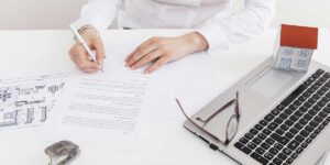 A professional fills out legal documents at a desk with architectural blueprints, a laptop, and a small house model.