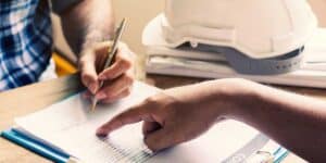 A person signs a written construction contract while another individual points out liability terms next to a white hardhat.